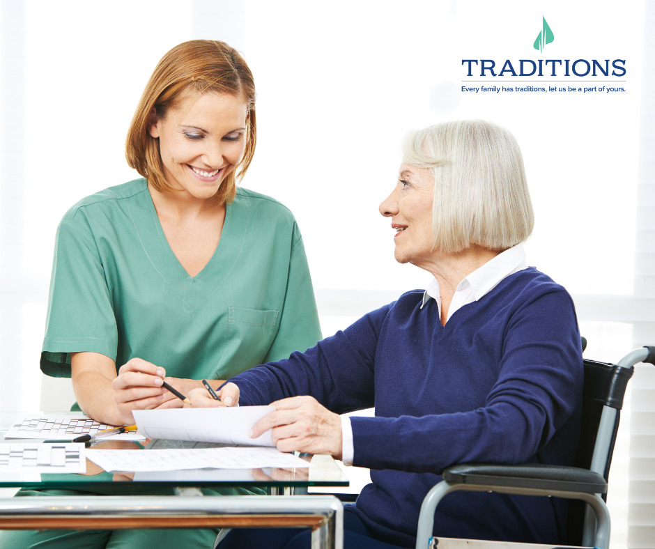 A nurse in green helps an elderly patient in indigo