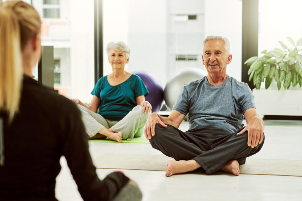 A senior man and senior woman sitting with their legs crossed on the ground in a yoga class looking at an instructor