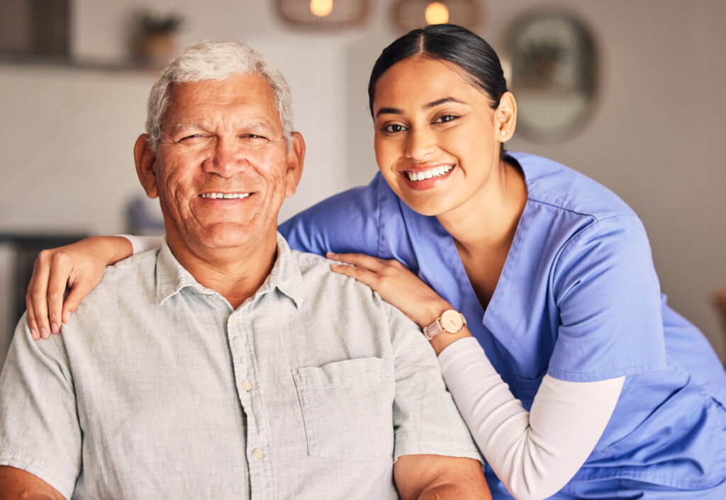 A male senior assisted living resident sitting as his female nursing assistant stand with her arm around him