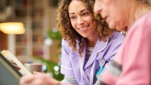 A female senior living employee in purple scrubs smiling while looking at a memory photo book with her memory care patient